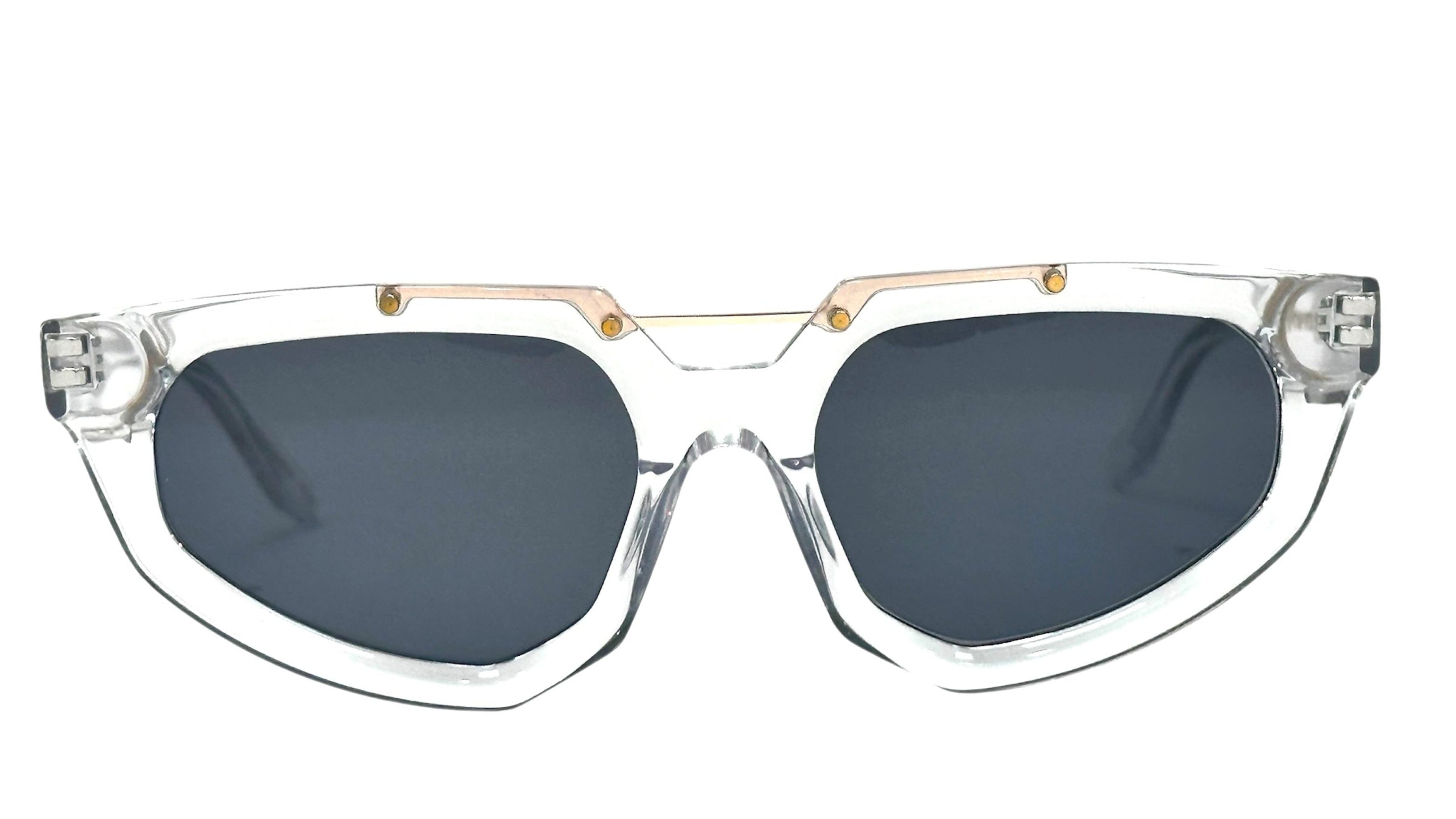 A pair of stylish sunglasses featuring a transparent frame and dark lenses. The design is angular with a unique shape, highlighted by gold accents at the bridge and temples.